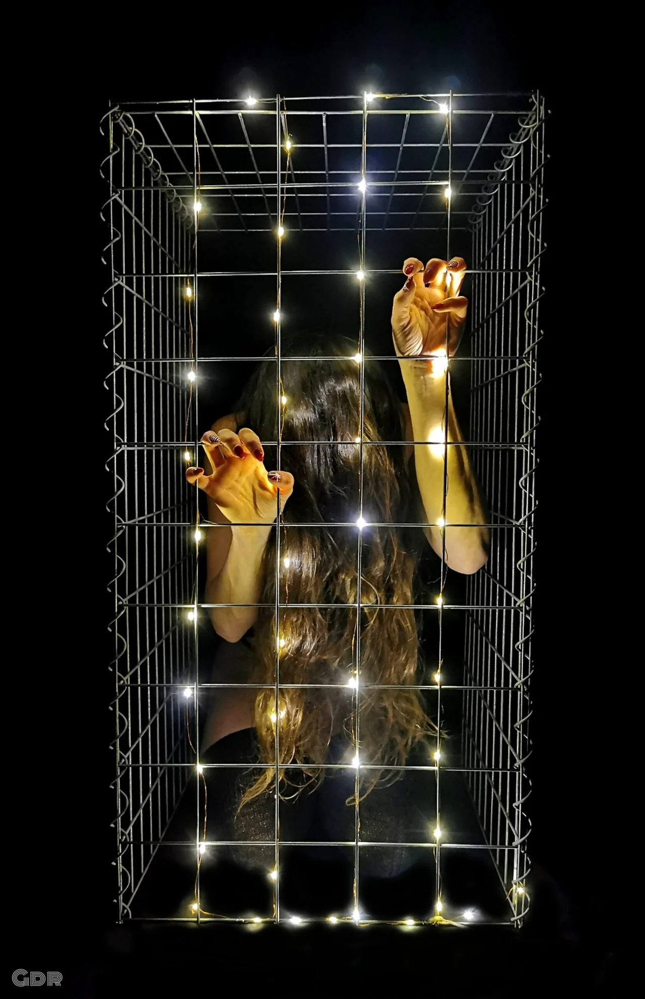 Femme enfermée dans une cage métallique éclairée de guirlandes lumineuses, symbolisant la tension entre lumière et enfermement.